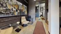 This single-story commercial space showcases a distinctive industrial-modern aesthetic that is immediately apparent upon entry. The front entrance features floor-to-ceiling glass panels flanking a branded glass door, flooding the reception area with natural light and offering views of the surrounding streetscape. Polished concrete floors extend throughout, complementing the raw texture of exposed concrete block walls and dark-painted surfaces.

The reception and lounge areas are anchored by reclaimed wood accent walls, against which large-scale abstract artwork is prominently displayed, adding bold visual interest. Seating includes a cream leather swivel chair and a rich dark leather sofa, creating a comfortable yet professional atmosphere. A glass-topped desk near the entry provides a functional workstation.

A central corridor, lined with pendant lighting, connects the front lounge to deeper interior spaces, which include additional rooms accessed through black-trimmed doors. Exposed ductwork near the ceiling reinforces the industrial character of the space. Carpeted utility areas near the rear transition zones offer contrast to the polished concrete flooring found elsewhere. Throughout, the design balances raw architectural elements with carefully curated furnishings and artwork to create a cohesive, contemporary environment.