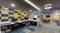 This single-floor space functions as a well-appointed business center with a thoughtfully designed interior that balances functionality and visual appeal. The workstation area features an L-shaped white desk along one wall, accommodating multiple computer setups complete with monitors, a printer, and ergonomic seating. The wall behind the workstations is adorned with bold, geometric acoustic panels in yellow, gray, and white, adding both a practical sound-dampening function and a striking visual element. Recessed and cove lighting along the ceiling provides warm, even illumination throughout the space. A decorative lattice-style window panel with an organic, cellular pattern serves as a stylish focal point, dividing the workstation area from the adjoining lounge. The flooring features a patchwork-style carpet in varied shades of blue and gray, adding texture and depth underfoot. Adjacent to the business center, a casual seating area with wooden chairs and tables offers a relaxed setting for meetings or informal gatherings. The overall ambiance is contemporary and energetic, making this space well-suited for both focused work and collaborative use.
