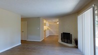 This single-floor space offers an open and functional layout that connects the living area to the kitchen, creating a cohesive flow throughout the interior. The living room is anchored by a striking corner fireplace set within a white-painted brick surround, serving as a standout architectural focal point that adds character and warmth to the space. A metal mesh screen completes the fireplace's classic look.

The flooring throughout is a warm wood-look material that lends continuity and visual warmth across the open plan. Neutral-toned walls provide a clean, versatile backdrop, while white baseboards and trim offer crisp contrast. The textured popcorn ceiling adds a traditional finish overhead.

The kitchen is visible through an open pass-through or transitional space, featuring white cabinetry and a ceiling fan, suggesting a practical and well-lit area suited for everyday use. A doorway off the living area provides access to additional rooms or storage.

Overall, the space presents a welcoming, straightforward interior with notable design elements like the brick fireplace that give it a distinctive and homey character.