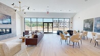 This contemporary single-floor community space offers a thoughtfully designed open-concept layout that seamlessly connects multiple functional areas. The main lounge is anchored by a dramatic floor-to-ceiling black-framed glass wall and entry doors, flooding the interior with abundant natural light and offering expansive outdoor views. A striking full-height tile-clad fireplace serves as the room's focal point, topped with a wall-mounted display and complemented by a designer sputnik chandelier that adds a touch of sophistication. Light hardwood flooring flows consistently throughout, unifying the space with warmth and continuity. Comfortable seating arrangements — including a leather sofa, upholstered accent chairs, and modular lounge seats — are thoughtfully positioned to encourage both conversation and relaxation. A dedicated dining area features a dark-topped table with sculptural cream chairs, positioned near a compact kitchenette outfitted with white cabinetry, open wood shelving, subway tile backsplash, stainless steel appliances, and a sink. Crisp white walls display carefully selected artwork, adding visual interest throughout. A secondary lounge nook offers additional flexible seating. The overall ambiance is clean, modern, and inviting, with high ceilings that enhance the sense of openness and volume across the entire space.