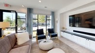 This thoughtfully designed game room offers a sophisticated blend of entertainment and comfort across a single, open floor. The centerpiece of the space is a striking built-in media wall finished in crisp white paneling, featuring a large wall-mounted flat-screen TV, an integrated soundbar, and a dark-topped cabinet system for ample storage. Flanking the media wall, gold-accented wall sconces add a warm, refined touch.

A dedicated wet bar area provides a functional yet elegant amenity, complete with a marble-veined stone countertop and backsplash, a stainless steel sink, a built-in beverage cooler, and dark wood cabinetry — ideal for entertaining.

The lounge area is anchored by a plush sofa and navy velvet accent chairs with gold-toned frames, arranged around a rounded white coffee table atop a textured area rug. Floor-to-ceiling black-framed windows and sliding glass doors flood the space with natural light and offer open views of the surrounding outdoor landscape, while breezy teal drapery adds a pop of color.

A separate dining nook features a solid wood table beneath a statement pendant light and is adorned with vibrant abstract artwork, making it equally suited for casual gatherings or game-day dining. Light wood-look flooring ties the entire space together seamlessly.