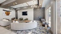 Located at 1550 East Campbell Avenue in Phoenix, AZ, this contemporary community space offers a sophisticated, single-floor layout designed for both socializing and relaxation. The open-plan kitchen and living area form the heart of the space, anchored by a large central island with a crisp white countertop and textured tile base, paired with sleek dark wood cabinetry and full stainless steel appliances. A striking sculptural pendant light fixture crowns the kitchen, complementing the soaring ceiling height above. The adjacent living area is defined by a sweeping curved sectional sofa, plush area rugs with abstract patterns, and a wall-mounted flat-screen TV set against geometric patterned wallcovering. Layered seating zones, including barrel chairs and lounge seating in rich earth tones, create an inviting and versatile atmosphere. A full shuffleboard table adds a recreational focal point to the space. Sputnik-style chandeliers provide warm ambient lighting throughout. A dedicated retreat nook features warm wood mosaic tile covering three walls, creating an intimate, cocoon-like atmosphere. Large windows and glass doors invite natural light, offering views to the outdoors and reinforcing the space's bright, modern character.