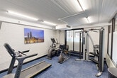 Located at 5021 S Valley Lane in Streamwood, IL, this fitness center offers a well-equipped exercise space designed to meet everyday workout needs. The room features white painted brick walls that lend a clean, modern aesthetic, complemented by blue modular carpet tiles that add a pop of color to the floor. Overhead fluorescent lighting ensures the space is consistently and evenly illuminated throughout.

The exercise room is outfitted with a range of cardio and strength training equipment, including a Matrix treadmill, an elliptical trainer, a recumbent bike, and a Hoist multi-station cable machine with an accompanying bench — providing a well-rounded selection for varied fitness routines. A wall-mounted flat-screen television is positioned near the treadmill area for entertainment during workouts.

A striking cityscape wall mural depicting the Chicago skyline at dusk serves as a vibrant focal point along one wall, adding visual interest to the space. Entry to the fitness area is through a sleek glass-panel partition with dark metal framing, creating a defined and polished transition point. Steps lead up to the entry vestibule, which features hardwood-style flooring. A wall-mounted control panel and a sanitation station are also present near the entrance, reflecting attention to cleanliness and user convenience.