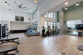 Located at 230 Bill Kennedy Way SE in Atlanta, GA, this well-appointed fitness center offers a comprehensive workout environment designed to accommodate a wide range of exercise needs. The facility is defined by its dramatic double-height ceilings and expansive floor-to-ceiling black-framed windows that flood the space with abundant natural light and provide views of the surrounding exterior. Light wood-tone flooring runs throughout, complementing the clean white walls and modern industrial aesthetic.

The cardio zone is generously equipped with multiple treadmills, elliptical cross-trainers, stair climbers, and rowing machines, all arranged to maximize the open, airy feel of the space. A dedicated cycling area features stationary bikes paired with a large flat-screen TV and a ballet-style barre along a full-length wall mirror, creating an immersive workout environment. The strength training section includes a comprehensive cable machine system, a multi-rack dumbbell station ranging across a wide variety of weights, adjustable benches, and functional training attachments such as battle ropes and resistance bands. Motivational wall art adds personality throughout. Large industrial ceiling fans and pendant lighting ensure the space remains comfortable and well-lit. Open storage cubbies along a textured brick accent wall provide practical organization for personal items.