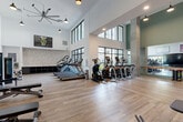 Located at 230 Bill Kennedy Way SE in Atlanta, GA, this well-appointed fitness center offers a comprehensive workout environment designed to accommodate a wide range of exercise needs. The facility is defined by its dramatic double-height ceilings and expansive floor-to-ceiling black-framed windows that flood the space with abundant natural light and provide views of the surrounding exterior. Light wood-tone flooring runs throughout, complementing the clean white walls and modern industrial aesthetic.

The cardio zone is generously equipped with multiple treadmills, elliptical cross-trainers, stair climbers, and rowing machines, all arranged to maximize the open, airy feel of the space. A dedicated cycling area features stationary bikes paired with a large flat-screen TV and a ballet-style barre along a full-length wall mirror, creating an immersive workout environment. The strength training section includes a comprehensive cable machine system, a multi-rack dumbbell station ranging across a wide variety of weights, adjustable benches, and functional training attachments such as battle ropes and resistance bands. Motivational wall art adds personality throughout. Large industrial ceiling fans and pendant lighting ensure the space remains comfortable and well-lit. Open storage cubbies along a textured brick accent wall provide practical organization for personal items.