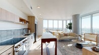 Located in the heart of Hollywood at 6390 De Longpre Ave, this elevated single-floor residence offers a sophisticated and light-filled living experience. The open-concept layout seamlessly connects the kitchen, dining, and living areas, all anchored by a dramatic curved wall of floor-to-ceiling windows that frame sweeping panoramic views of the Los Angeles skyline, including a glimpse of the iconic Netflix sign.

The kitchen is outfitted with light wood cabinetry, open shelving, a blue tile backsplash, stainless steel appliances, and a generous marble-topped island with a wood base that doubles as a dining surface. Wide-plank hardwood flooring flows throughout the main living area, complemented by a striking exposed concrete cylindrical column that adds an industrial architectural accent.

The living area is bathed in natural light and opens to an expansive city view, creating an airy, open ambiance. The bedroom features large picture windows with equally impressive cityscape views, soft neutral tones, and a distinctive concrete-panel accent wall with driftwood art.

Two bathrooms serve the residence — one with marble-look tile flooring, a wood-vanity, and a glass-enclosed shower, and another featuring full-height tile, a glass shower enclosure, and warm accent lighting, both finished with refined, modern detailing throughout.
