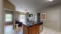 This single-floor unit offers a thoughtfully designed open-concept layout that seamlessly connects the kitchen, dining, and living areas. The kitchen is a functional centerpiece, featuring a large island with a double-basin sink, dark countertops, warm wood cabinetry, stainless steel appliances including a refrigerator, microwave, and range, and bar-height seating for casual dining. Tile flooring runs throughout the kitchen and entry areas, while warm hardwood flooring extends into the living spaces and bedrooms.

The living room is bright and inviting, benefiting from a sliding glass door that opens to a private balcony and allows generous natural light to fill the space. A ceiling fan adds comfort, and a wall-mounted TV is included. Built-in shelving provides additional display and storage options nearby.

The unit includes two bedrooms, each featuring hardwood floors, ceiling fans, and windows with blinds that frame views of surrounding greenery. One bedroom includes its own exterior door access to a private outdoor area.

Two full bathrooms serve the unit, each finished with tile flooring, dark countertops, wood cabinetry, and generous storage. One bathroom features a tub-shower combination, while the other includes a closet for added convenience. The overall aesthetic is clean, neutral, and functional throughout.