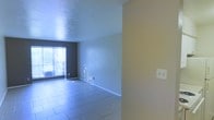 This single-floor space presents an open, unfurnished interior characterized by light tile flooring that extends throughout the main living area. The layout is straightforward, with a clear sightline from the entry point toward a large window at the far end, which allows natural light to filter into the room and cast a bright, airy quality across the space. The walls are painted in neutral tones — a soft blue-gray on the main living area walls and a warm beige on the adjacent partition wall — creating a simple, versatile backdrop suitable for a variety of uses. A textured ceiling adds subtle visual interest overhead. White baseboards run along the perimeter, providing a clean finish to the walls and flooring junction. Electrical outlets are visible along the walls, indicating practical utility placement throughout. The open floor plan allows for flexible furniture arrangement, and the absence of interior obstructions contributes to a sense of openness. The large window at the end of the room serves as the primary source of natural light, brightening the interior and offering a connection to the outside environment.
