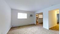 This single-floor space features a clean, straightforward layout designed for functional living. The main living area is finished with wall-to-wall carpet in a neutral tone, lending a soft, understated feel to the room. White walls throughout create a bright, open atmosphere, complemented by a window that allows natural light to filter in. A wall-mounted air conditioning unit and a baseboard heating system ensure climate control throughout the seasons.

An open doorway connects the living area directly to the kitchen, which is visible from the main room and features white appliances, including a range and what appears to be a refrigerator. The kitchen cabinetry adds a warm wood-toned contrast against the lighter finishes of the space. The transition between rooms is seamless, contributing to an easy flow within the layout.

The space maintains a minimal, utilitarian aesthetic with clean lines and a neutral color palette. The combination of carpet in the living area and the practical kitchen setup makes this a well-organized, easy-to-maintain space suited for everyday living.