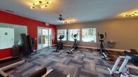 Located at 1821 Buckeye Dr in Richmond Hill, Georgia, this fitness center is a well-appointed, single-floor facility designed to support a comprehensive workout experience. The space is defined by bold red accent walls contrasted against neutral gray tones, creating an energizing atmosphere that motivates exercise.

The fitness floor is generously sized and covered in durable, patterned carpet tiles that add texture and visual interest throughout. Multiple large windows line the walls, flooding the space with natural light and offering views of the surrounding outdoor area, including what appears to be a pool and green landscaping beyond.

The facility is equipped with a full range of cardio machines, including treadmills, ellipticals, and stationary bikes, all arranged to allow comfortable movement between stations. A dedicated free weight area features a multi-tiered dumbbell rack alongside kettlebells, catering to strength training needs. A cable and multi-function weight machine rounds out the strength equipment offerings.

Overhead, a textured ceiling hosts a combination of ceiling fans and stylish spur-light fixtures, ensuring proper air circulation and ample illumination throughout. Glass-panel doors provide direct access to the outdoor area, further connecting the interior to the surrounding community. The overall ambiance is clean, functional, and inviting for users of all fitness levels.