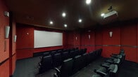 Located at 4950 E Van Buren St in Phoenix, AZ, this single-floor private screening room delivers an immersive cinematic experience through a carefully curated interior design. The room is defined by its bold, deep-red walls that extend from floor to ceiling, creating a dramatic and intimate atmosphere reminiscent of a classic movie theater. A continuous dark horizontal trim band runs along the perimeter of the walls, adding a refined architectural detail that separates the upper and lower wall sections.

The screening room is equipped with a large, centrally positioned projection screen mounted at the front wall, paired with a ceiling-mounted projector at the rear of the room. Multiple rows of black leather theater-style seats with integrated armrests and cup holders are arranged in a tiered layout, ensuring clear sightlines from every position. Recessed ceiling lighting and wall-mounted sconces provide subtle, ambient illumination throughout the space.

Decorative touches enhance the cinematic theme, including a series of framed artwork pieces displayed along the rear wall and large vintage film reel sculptures mounted near the exit corridor, reinforcing the entertainment-focused identity of the space. A side console table provides a functional surface for equipment. The dark carpeted flooring and dark ceiling complete the immersive, light-controlled environment purpose-built for high-quality film viewing.
