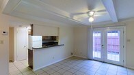 Located on Spring Hill Avenue in Mobile, Alabama, this two-story property offers a functional and straightforward layout designed for comfortable everyday living. The main living area features light ceramic tile flooring and a textured ceiling with an integrated ceiling fan, providing both comfort and airflow. Exposed beam detailing adds a subtle architectural character to the space. A pass-through kitchen counter with dark countertops connects the kitchen to the living and dining area, creating an open, connected feel. The kitchen includes dark cabinetry and essential appliances visible from the pass-through. White wainscoting along the lower portion of the kitchen partition adds a classic touch to the interior. Natural light fills the main living space through a full-length glass door that opens to an outdoor area, blurring the line between interior and exterior living. The door is framed with white curtains, enhancing the bright, airy atmosphere of the room. The two-story configuration provides additional living or sleeping space on the upper level, adding versatility to the overall floor plan. The property's location on Spring Hill Avenue places it in one of Mobile's well-known corridors, offering convenient access to local amenities and services.