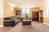 Located at 165 E Grand Ave in Rahway, NJ, this single-floor space is designed as a welcoming communal environment with a cohesive and vibrant interior aesthetic. The layout is anchored by a comfortable lounge area furnished with upholstered sofas, decorative throw pillows, and a wooden coffee table accented by a floral arrangement. Large floor-to-ceiling windows flood the seating area with natural light while offering open views of the surrounding greenery, creating an inviting and relaxed atmosphere.

The walls throughout are finished in a warm golden yellow with crisp white crown molding and chair-rail detailing, complemented by bold red accent doors that add a striking visual contrast. Decorative framed artwork and mirrors are thoughtfully placed along the walls, contributing to the space's polished character. The floors are covered in a textured, muted rose-toned carpet that ties the color palette together.

A separate dining nook, accessible from the main lounge, features a dark wood table and chairs set beside large windows that provide views of the neighborhood streetscape. Wall sconces offer soft supplemental lighting throughout. The overall ambiance is warm, organized, and purposefully designed for communal comfort and everyday use.