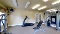 This fitness room offers a functional and well-organized space designed to support a variety of workout routines. The room features neutral beige walls and carpeted flooring, creating a clean and comfortable environment for exercise. Overhead, a series of bright fluorescent lighting fixtures ensure the space is thoroughly illuminated throughout the day.

Key equipment includes a treadmill for cardiovascular training, an adjustable workout bench for strength and flexibility exercises, and a cable machine anchored along one wall, which supports a wide range of resistance training movements. A large wall-mounted mirror runs along one side of the room, enhancing the sense of space while allowing users to monitor their form during workouts.

The room is accessed through elegant French doors with glass panes, adding a refined architectural touch to the otherwise utilitarian space. A standard interior door is also visible, suggesting additional access or an adjoining room. Ventilation is provided through ceiling vents, and safety is addressed with clearly marked exit signage. Overall, the space delivers a practical and accessible fitness environment.