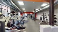 This fitness center offers a spacious, single-floor layout designed to accommodate a wide range of workout routines. The space features an industrial aesthetic, highlighted by exposed ductwork running along the ceiling, dark structural columns, and bold orange accent walls that energize the environment. Rubber flooring throughout provides both safety and durability across all exercise zones.

The cardio section is well-appointed with a row of treadmills, elliptical machines, and stair climbers, several of which are paired with wall-mounted flat-screen televisions for entertainment during workouts. Large mirrors line portions of the walls, enhancing the sense of openness and allowing users to monitor their form.

The strength training area includes a power rack with weight plates, a cable machine, a adjustable bench, and a selection of dumbbells neatly organized on a rack. Stability balls and wall-mounted fitness pads round out the functional training options. A freestanding heavy punching bag adds a combat training element to the space.

Recessed panel lighting ensures the entire floor is evenly and brightly illuminated. Windows along the walls allow natural light to filter in, while a high ceiling with exposed rafters in the main area adds a sense of volume and openness. A water station is conveniently accessible within the facility.