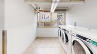 Located at 221 Queen St S in Kitchener, Ontario, this shared laundry facility is a practical and efficiently organized space designed to accommodate high-volume use. The room is finished with neutral, light-toned ceramic floor tiles throughout, contributing to a clean and bright atmosphere. Exposed ductwork and overhead fluorescent lighting run along the white ceiling, ensuring the space is well-lit at all times.

The laundry area is equipped with a row of front-loading washing machines and a combination of stacked and side-by-side dryer units, providing ample capacity for multiple users simultaneously. A large central folding table offers a convenient surface for sorting and folding laundry. A utility sink is tucked into a dedicated nook, and a storage cabinet provides additional organizational space.

Natural light filters in through windows along one wall, offering views of the surrounding neighbourhood. Access control is managed via a keypad entry system, enhancing security. The utility room houses building infrastructure, including visible piping and mechanical systems. A laundry cart, waste bin, and wall-mounted artwork add functional and aesthetic touches. The overall layout is straightforward and user-friendly, making this a highly practical shared amenity.