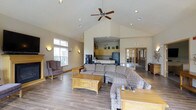 This single-floor community facility offers a comprehensive range of amenities designed for comfort and active living. The grand entrance is framed by tall white columns beneath a covered portico, creating a welcoming first impression. Inside, the main lounge area impresses with soaring vaulted ceilings, recessed lighting, and a ceiling fan that adds to the airy atmosphere. Wood-look flooring runs throughout, complementing the warm, neutral tones of the walls. The lounge is furnished with generous sectional seating, accent chairs, and a wooden coffee table, all centered around an adjoining kitchenette equipped with cabinetry and a full-size refrigerator. A dedicated fitness center provides a well-equipped workout space featuring treadmills, a cable machine, free weights, and mirrored walls that enhance the sense of space. The indoor hot tub room offers a tranquil retreat, surrounded by tiled walls, large windows overlooking the outdoor pool area, and bench seating. Outdoors, a generously sized swimming pool is lined with lounge chairs and anchored by sweeping views of lush, rolling green hills and mature trees. A pergola-covered seating area with bench seating provides a shaded spot to enjoy the natural surroundings, completing this thoughtfully designed community space.