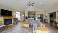 This single-floor community facility offers a comprehensive range of amenities designed for comfort and active living. The grand entrance is framed by tall white columns beneath a covered portico, creating a welcoming first impression. Inside, the main lounge area impresses with soaring vaulted ceilings, recessed lighting, and a ceiling fan that adds to the airy atmosphere. Wood-look flooring runs throughout, complementing the warm, neutral tones of the walls. The lounge is furnished with generous sectional seating, accent chairs, and a wooden coffee table, all centered around an adjoining kitchenette equipped with cabinetry and a full-size refrigerator. A dedicated fitness center provides a well-equipped workout space featuring treadmills, a cable machine, free weights, and mirrored walls that enhance the sense of space. The indoor hot tub room offers a tranquil retreat, surrounded by tiled walls, large windows overlooking the outdoor pool area, and bench seating. Outdoors, a generously sized swimming pool is lined with lounge chairs and anchored by sweeping views of lush, rolling green hills and mature trees. A pergola-covered seating area with bench seating provides a shaded spot to enjoy the natural surroundings, completing this thoughtfully designed community space.