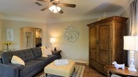 This single-floor residence offers a comfortable and well-appointed living environment with a warm, welcoming ambiance. The living room serves as the heart of the home, showcasing rich hardwood-style flooring, neutral-toned walls, and elegant crown molding that adds a refined architectural touch. A ceiling fan with a built-in light fixture provides both comfort and illumination, complementing the warm glow of accent lamps. The space is furnished with a plush, tufted gray sofa and a classic tufted ottoman, creating an inviting seating arrangement centered around a decorative area rug. A large, handsome wood armoire anchors one wall and adds a sense of traditional craftsmanship to the room. A circular ornamental wall piece serves as a focal point, adding artistic character to the neutral backdrop. Sightlines from the living area reveal a connected kitchen space, suggesting an open and functional floor plan that promotes easy flow between living and entertaining areas. The overall aesthetic of this home blends classic design elements with cozy, lived-in comfort, making it a practical and visually appealing single-floor space.