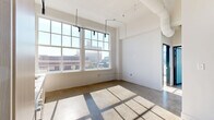 Located in St. Louis, MO, this two-bedroom, one-bathroom loft on Locust Street delivers an urban industrial aesthetic with thoughtful modern updates. The open-plan living and kitchen area is anchored by floor-to-ceiling grid windows that flood the space with natural light and frame sweeping city views. Polished concrete floors run throughout, complementing the loft's industrial character, while soaring ceilings create an airy, expansive feel well beyond the unit's modest footprint. The kitchen features sleek light-wood cabinetry, stainless steel appliances including a built-in oven and microwave, and a clean countertop workspace beneath a striking black track lighting system. A standout design element is the oversized frosted sliding glass partition, which separates spaces with a contemporary flair. The primary bedroom benefits from large windows overlooking the surrounding cityscape. The bathroom continues the modern industrial theme with dark accent walls, a glass-enclosed shower, and wall-mounted fixtures. A hallway connects the rooms efficiently, maximizing the use of the overall layout. This loft combines raw architectural character with refined finishes, making it a compelling urban living space in the heart of St. Louis.