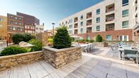 Located in the heart of Wilmington, Delaware, this outdoor amenity space offers a well-appointed urban retreat set against the backdrop of a modern multi-story building with a contemporary façade combining light panel cladding and classic brick. The centerpiece of the space is a generously sized swimming pool with crystal-clear water, surrounded by a wood-toned deck lined with yellow lounge chairs, ideal for sunbathing and relaxation. A handsome wooden pergola provides a shaded seating area adjacent to the pool, adding architectural character and comfort to the sun deck. Lush landscaping, including mature shrubs and greenery, frames the perimeter, softening the urban environment and enhancing the sense of a private oasis. Stone retaining walls with natural stacked-stone detailing add a refined, textural element throughout the grounds. A gated walkway connects the pool area to the building entrance, ensuring easy access while maintaining security. On the patio side, large format stone pavers create a generous outdoor dining area furnished with multiple tables and seating for groups of all sizes, making it well-suited for social gatherings. Large glass doors and windows along the brick base of the building allow natural light to flow between the interior and exterior spaces, reinforcing a seamless indoor-outdoor connection throughout this thoughtfully designed amenity level.