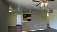 This single-floor residence offers a clean, open-concept layout designed for comfortable everyday living. The main living area is finished with rich, dark hardwood flooring that extends throughout, providing a warm and cohesive look. A ceiling fan with integrated lighting fixtures is centrally positioned overhead, ensuring consistent airflow and ambient light across the space.

The kitchen is defined by a raised pass-through counter that opens directly to the living area, creating a connected and social atmosphere. Dark cabinetry lines the kitchen walls, complemented by black appliances and a countertop with a built-in sink beneath an under-cabinet range hood. This contrasting combination of dark tones gives the kitchen a bold, contemporary character.

Natural light enters the space through a glass-paneled exterior door, which provides access to an outdoor area while brightening the interior. The neutral, taupe-toned walls throughout offer a versatile backdrop well-suited to a variety of furnishing styles. White baseboards provide a crisp finishing detail that ties the overall aesthetic together.

The layout is efficient and well-organized, with a clear visual flow between the living and kitchen areas. The overall ambiance is modern and understated, combining functional design elements with a cohesive color palette.