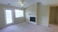 This single-story living space offers a straightforward and functional layout with a warm, neutral aesthetic. The main living area is finished with wall-to-wall carpet in a soft beige tone, contributing to a cozy and comfortable feel underfoot. Walls are painted in a light, warm neutral that reflects natural light effectively throughout the space.

A built-in fireplace serves as the focal point of the living area, framed by a clean white mantel and set against an angled accent wall that adds subtle architectural interest. The fireplace features a classic black surround, providing a traditional yet understated design element.

Natural light enters the room through a window fitted with horizontal blinds, allowing for adjustable light control. A ceiling fan with an integrated light fixture is centrally positioned, providing both air circulation and overhead illumination to the space.

The room benefits from clean lines and a simple design approach, making it a versatile canvas for a variety of furniture arrangements and décor styles. Baseboards and trim throughout are finished in white, complementing the neutral palette. The overall ambiance is quiet and move-in ready.