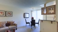 This single-floor space offers a thoughtfully arranged open layout that combines living and dining areas into a cohesive, functional environment. The dining area serves as a central feature, accommodating a round glass-top table surrounded by dark-framed chairs beneath a pendant light fixture that draws the eye upward toward the textured ceiling. A decorative square mirror with a bold striped frame adorns the wall, adding visual interest and depth to the space. Large vertical-blind-covered sliding glass doors flood the dining area with abundant natural light and provide direct access to an outdoor balcony, offering views beyond the interior. Adjacent to the dining space, a partial view of the living area reveals a cozy seating arrangement with a light-toned sofa and accent pillow. To the right, a kitchen counter with dark cabinetry overhead transitions smoothly into the open-plan layout, creating a natural boundary between cooking and living zones. The space is finished with neutral-toned carpeting throughout, contributing to a warm and understated ambiance that ties each area together cohesively.