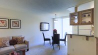 This single-floor space offers a thoughtfully arranged open layout that combines living and dining areas into a cohesive, functional environment. The dining area serves as a central feature, accommodating a round glass-top table surrounded by dark-framed chairs beneath a pendant light fixture that draws the eye upward toward the textured ceiling. A decorative square mirror with a bold striped frame adorns the wall, adding visual interest and depth to the space. Large vertical-blind-covered sliding glass doors flood the dining area with abundant natural light and provide direct access to an outdoor balcony, offering views beyond the interior. Adjacent to the dining space, a partial view of the living area reveals a cozy seating arrangement with a light-toned sofa and accent pillow. To the right, a kitchen counter with dark cabinetry overhead transitions smoothly into the open-plan layout, creating a natural boundary between cooking and living zones. The space is finished with neutral-toned carpeting throughout, contributing to a warm and understated ambiance that ties each area together cohesively.