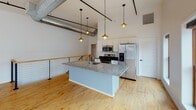 Located at 1255 University Ave in Rochester, NY, this two-floor loft-style space masterfully combines industrial character with contemporary design. The main level opens into a generous, light-filled open floor plan showcasing warm hardwood floors, soaring ceilings with exposed beams, and visible ductwork that pays homage to the building's industrial heritage. Large steel-framed grid windows flood the space with natural light while offering outdoor views, creating a bright and airy atmosphere throughout.

The well-appointed kitchen features white cabinetry, a granite-topped island with a sink, stainless steel appliances including a refrigerator, stove, and built-in microwave, all illuminated by stylish pendant lighting. A dramatic open-riser metal staircase connects the two floors, serving as both a functional and architectural focal point.

The second level continues the open loft aesthetic with additional living or working space, pendant lighting, and windows that bring in natural light. Multiple rooms across both floors provide flexibility, each finished with the same hardwood flooring. Two bathrooms are present — one featuring a granite vanity, tiled shower, and tile flooring, and another spacious bathroom with large-format tile. The overall ambiance is modern, urban, and versatile.