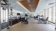 Located at 170 S 6th St in Philadelphia's historic Old City neighborhood, this fitness center is a thoughtfully designed, single-floor facility offering a comprehensive workout environment. The main exercise floor is spacious and open, featuring a distinctive coffered ceiling with warm wood paneling and integrated ambient lighting that lends a polished, upscale aesthetic. Recessed lighting and large perimeter windows allow natural light to fill the space, offering views of the surrounding cityscape — including glimpses of notable Philadelphia architecture — while users work out on the cardio equipment lining the window-facing wall.

The main floor is outfitted with a broad selection of modern machines, including treadmills, ellipticals, and cycling equipment, alongside a dedicated strength training area featuring cable machines, weight benches, and a full dumbbell rack. Large mirrors along one wall enhance the sense of space and support proper form during workouts.

A separate studio room provides a more intimate setting for group fitness or individual training. Its hardwood-style flooring, deep teal accent walls, floor-to-ceiling mirrors, wall-mounted barre, and collection of stability balls and free weights make it well-suited for yoga, pilates, barre, or stretching. An entry corridor with built-in cabinetry and a water station connects the two areas seamlessly.