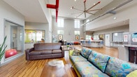 Located at 258 University Dr in Marion, Ohio, this single-floor community space is designed with an open, flexible layout that encourages social interaction and active engagement. The architecture is immediately striking, with dramatically high ceilings that amplify the sense of spaciousness throughout the main common area. Exposed red ductwork and an oversized industrial ceiling fan add a modern, urban character to the space.

The main gathering area features warm hardwood-style flooring and is furnished with a mix of seating options, including leather sofas, colorful patterned couches, and vibrant hexagonal ottomans in bold primary colors. A large wall-mounted flat-screen television anchors one end of the lounge, complemented by stylish media furniture and decorative greenery.

The kitchen area includes a stainless steel island with bar-height seating, stainless appliances, and a ventilation hood, making it well-suited for communal use. Natural light pours in through clerestory windows positioned high on the walls, brightening the interior throughout the day. Large ground-level windows offer views of the surrounding outdoor greenery.

A dedicated fitness area is visible within the space, equipped with exercise equipment. The overall ambiance is lively and welcoming, with colorful accents, curated artwork, and thoughtful furnishings creating an energetic yet comfortable environment.