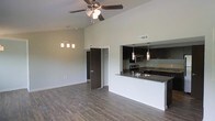 This single-floor residence showcases a thoughtfully designed open-concept layout that maximizes both space and functionality. The living area is defined by impressively high vaulted ceilings that lend an airy, expansive feel to the interior. A ceiling fan with a light fixture is centrally positioned overhead, complementing an elegant pendant chandelier that adds a warm, decorative touch to the space. Durable hardwood-style flooring runs throughout, providing a cohesive and contemporary aesthetic.

The kitchen is seamlessly integrated into the living area via a peninsula-style breakfast bar topped with dark granite countertops, creating a natural gathering point. Dark espresso cabinetry lines the kitchen walls, offering generous storage, while stainless steel appliances — including a range and microwave — add a modern, polished finish. Pendant lighting above the bar enhances both the ambiance and practicality of the cooking and dining space.

The open layout connects naturally to additional areas of the residence, with doorways leading to further rooms beyond. Neutral wall tones throughout keep the atmosphere clean and versatile, making the space easy to personalize. Overall, the residence strikes a balance between modern design elements and comfortable, livable features.