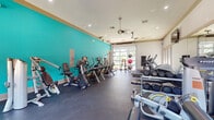This fitness center is a spacious, single-floor facility designed to support a comprehensive workout experience. The interior is immediately distinguished by a bold teal accent wall that adds a vibrant, energizing atmosphere to the space, complemented by neutral tones on the remaining walls and a clean white ceiling with recessed lighting and a ceiling fan for comfort during workouts.

The flooring throughout is durable dark rubber matting, well-suited for heavy equipment and high-activity use. The facility is generously outfitted with a wide variety of cardio machines, including treadmills, ellipticals, recumbent bikes, and upright cycles, arranged to allow comfortable circulation. A dedicated strength training area features a full dumbbell rack, weight benches, a cable machine system, and a Hoist multi-functional trainer, catering to users across all fitness levels.

Natural light pours in through multiple large windows and a full-width garage-style glass panel door, offering views to the outdoors and creating an open, airy feel. Wall-mounted flat-screen televisions are positioned for easy viewing from multiple workout stations. Full-length mirrors along one wall enhance the sense of space and allow for form-checking during exercise. Medicine balls and stability equipment add further versatility to the training options available in this well-appointed fitness facility.