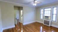This single-floor space offers a clean, classic interior defined by gleaming hardwood floors that reflect natural light throughout the room. The ceiling is adorned with crisp crown molding, adding a traditional architectural detail that lends the space a polished, timeless character. A ceiling fan with an integrated light fixture is centrally mounted, providing both air circulation and ambient lighting.

The walls are finished in a neutral, warm beige tone, creating a bright and versatile backdrop. A window fitted with blinds on one side allows natural light to filter in while offering privacy. A wall-mounted radiator beneath the window suggests a traditional heating system built into the structure.

An open doorway leads to what appears to be a bathroom, which is visible with its white fixtures and tiled flooring. An adjacent door provides access to an additional room or hallway beyond, suggesting a connected, functional layout. Wide white baseboards frame the perimeter of the room, complementing the crown molding and contributing to the overall sense of classic detailing. The overall ambiance is light, airy, and well-maintained, with traditional architectural features throughout.