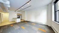 This single-floor space offers an open-concept layout that seamlessly integrates a living area with a functional kitchen. The main living area is finished with a distinctive patterned carpet featuring a grey and orange tile design, adding a contemporary visual element to the floor. Clean white walls and a smooth white ceiling contribute to a bright and airy ambiance throughout.

The kitchen is tucked neatly to one side, equipped with wood-toned cabinetry, stainless steel appliances including a refrigerator and microwave, and a compact kitchen island or peninsula with a dark countertop that serves as both a workspace and a natural divider between the kitchen and the open living space. A pendant light hangs above the kitchen area, providing focused illumination.

The main living area is illuminated by a bold track lighting system mounted on the ceiling, offering flexible and adjustable lighting across the space. A baseboard heating system runs along the lower portion of one wall, ensuring practical climate control throughout. The open floor plan and minimal design aesthetic make this a versatile space well-suited for a variety of uses, from residential living to a studio or creative workspace.
