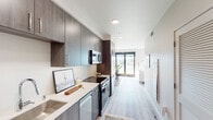 This well-appointed single-floor residence showcases a thoughtfully designed contemporary interior with a cohesive aesthetic carried throughout each space. The galley-style kitchen is a standout feature, outfitted with dark wood-grain cabinetry extending to the ceiling for ample storage, crisp white quartz countertops, a stainless steel sink, and a full suite of stainless steel appliances including an electric range, over-the-range microwave, and dishwasher. Light wide-plank flooring runs continuously from the kitchen through to the living area, enhancing the sense of flow and space. The living room is bright and inviting, benefiting from multiple windows and a full glass sliding door that opens to an outdoor area, flooding the room with natural light and offering views of the surrounding neighborhood. Built-in cabinetry with a media console adds both function and style to the living space. The bedroom is designed for comfort and efficiency, featuring overhead cabinetry built directly above the bed for smart storage, complemented by warm ambient lighting. The bathroom is clean and minimal, featuring a full soaking tub with shower and white tile surround, finished with polished chrome fixtures. Throughout the residence, white walls, recessed and flush-mount lighting, and louvered closet doors contribute to a modern, airy atmosphere.