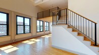 This thoughtfully designed two-level studio offers an open, airy layout defined by soaring ceilings, warm hardwood floors, and an abundance of natural light. The main living area is anchored by a striking wall of large, black-framed windows that flood the space with sunlight and frame views of the surrounding cityscape. A beautifully crafted staircase with warm wood treads and sleek black metal railings leads to the lofted sleeping area above, which features the same rich hardwood flooring, a natural wood plank ceiling, and its own set of large windows overlooking the city. The loft also includes a dedicated walk-in closet with built-in shelving and hanging rods for ample storage. The kitchen is well-appointed with light wood cabinetry, a tile backsplash, recessed lighting, and a full suite of white appliances including a gas range, dishwasher, and refrigerator. The bathroom is elegantly finished with white penny-tile flooring, a classic pedestal sink, a tile accent border, and a separate water closet. A stacked washer and dryer unit is conveniently integrated within the bathroom area. Throughout the space, the combination of industrial-inspired details and warm natural materials creates a clean, urban aesthetic.