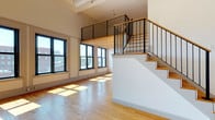 This thoughtfully designed two-level studio offers an open, airy layout defined by soaring ceilings, warm hardwood floors, and an abundance of natural light. The main living area is anchored by a striking wall of large, black-framed windows that flood the space with sunlight and frame views of the surrounding cityscape. A beautifully crafted staircase with warm wood treads and sleek black metal railings leads to the lofted sleeping area above, which features the same rich hardwood flooring, a natural wood plank ceiling, and its own set of large windows overlooking the city. The loft also includes a dedicated walk-in closet with built-in shelving and hanging rods for ample storage. The kitchen is well-appointed with light wood cabinetry, a tile backsplash, recessed lighting, and a full suite of white appliances including a gas range, dishwasher, and refrigerator. The bathroom is elegantly finished with white penny-tile flooring, a classic pedestal sink, a tile accent border, and a separate water closet. A stacked washer and dryer unit is conveniently integrated within the bathroom area. Throughout the space, the combination of industrial-inspired details and warm natural materials creates a clean, urban aesthetic.