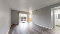 This single-floor space is designed with a clean, contemporary aesthetic throughout. The main living area is open and airy, finished with wood-style laminate flooring in a cool gray tone and neutral gray walls that create a cohesive, modern look. A textured popcorn ceiling adds dimension overhead, while a large sliding glass door fills the space with natural light and provides direct access to a private outdoor balcony, offering views of the exterior surroundings.

Adjacent to the living area, a secondary room features its own window with blinds, built-in open shelving, and a closet, making it a versatile space suitable for use as a bedroom or home office. The flooring continues seamlessly throughout, reinforcing the unit's consistent design language.

The galley-style kitchen is efficiently laid out with white cabinetry, marble-look countertops, a white subway tile backsplash, and stainless steel appliances including a range, microwave, and refrigerator. Light wood-tone flooring brightens the compact cooking space.

The bathroom is well-appointed with a white vanity topped with a marble-look surface, a soaking tub, and full-height marble-style tile surrounding the tub and shower area, lending an elevated, polished feel to the space.