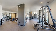 Located at 3200 Pleasant Grove Blvd in Roseville, CA, this fitness center offers a generously sized, single-floor layout designed to accommodate a full range of workout styles and fitness goals. The main exercise area is bright and open, featuring high white ceilings with recessed lighting and striking arched windows and glass doors that flood the space with natural light while offering views of the surrounding outdoor environment. Durable speckled rubber flooring covers the main floor, providing both safety and comfort underfoot. The exercise floor is comprehensively outfitted with a wide array of strength and cardio equipment, including cable machines, selectorized weight stations, leg press machines, treadmills, rowing machines, ellipticals, and a full rack of dumbbells. Wall-mounted flat-screen televisions and large mirrors enhance the workout experience throughout the space. A separate functional training room, finished with warm wood-look flooring, provides a dedicated area for dynamic training and features kettlebells, medicine balls, resistance bands, plyo boxes, and foam mats organized on a storage rack. The clean, modern aesthetic is carried throughout both areas, with crisp white walls, dark-trimmed doors, and a layout that maximizes usable floor space while maintaining an open, motivating atmosphere.