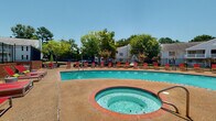 Bradford Place Pool, located in Byram, Mississippi, is a well-appointed outdoor aquatic amenity designed for relaxation and recreation. The centerpiece of the space is a large, curved swimming pool with clear turquoise water, bordered by a brick-trimmed edge that adds a classic, polished aesthetic. Adjacent to the main pool sits a circular hot tub, also framed in brick, providing a dedicated space for hydrotherapy and leisure.

The expansive pool deck is finished in a textured aggregate surface, offering both visual appeal and slip resistance. The deck is generously furnished with vibrant red chaise lounge chairs accented with green cushions, arranged to maximize sun exposure throughout the day. Patio tables with umbrellas are also available, providing shaded seating options.

The entire pool area is enclosed by a black wrought iron fence, ensuring both safety and security while maintaining an open, airy feel. Mature trees and flowering shrubs surround the perimeter, adding natural greenery and color to the environment. A community building is situated at one end of the deck, and a sports court with lighting is visible nearby. The open sky above and lush surroundings create a resort-like atmosphere, making this pool area a standout communal feature of the Bradford Place community.