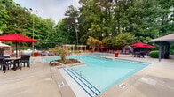 Located at 1600 Winters Creek Dr in Atlanta, Georgia, this single-level outdoor amenity facility offers a well-appointed pool complex set against a backdrop of mature, towering trees that provide natural privacy and shade. The centerpiece of the space is a dual-pool layout, featuring two generously sized swimming pools with clear blue water, blue-striped lane markings, and entry handrails for easy access. Surrounding the pools is an expansive concrete pool deck furnished with rows of dark lounge chairs and striking red patio umbrellas, creating a vibrant and cohesive aesthetic throughout the space. A covered pavilion runs along one side of the facility, supported by dark structural columns and featuring overhead lighting, providing a shaded retreat for lounging or dining. Outdoor dining tables and chairs are arranged beneath the pavilion and along the deck, ideal for social gatherings. Decorative landscaping, including flowering plants and ornamental shrubs, adds color and texture between the pool areas. A pergola structure near the rear of the pool deck provides an additional shaded seating area. The main building, finished in gray stucco with a pitched shingle roof and bold red accent doors, houses support facilities and storage. The entire complex is enclosed by fencing, ensuring a secure and serene environment for residents.