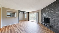 Located in Kansas City, MO, this well-appointed 2-bedroom, 2-bathroom residence offers a comfortable and functional single-floor layout. The open living room is a standout space, featuring light hardwood-style flooring, a striking dark stone accent fireplace, exposed dark wood beam details, and a sliding glass door that leads to a private balcony overlooking lush greenery. A pass-through window connects the living area to the kitchen, enhancing the open feel. The kitchen is outfitted with white cabinetry accented by black hardware, dark granite-style countertops, a subway tile backsplash, stainless steel appliances including a dishwasher and refrigerator, and a black matte faucet. Both bedrooms are carpeted and include ceiling fans with lighting. The primary bedroom features a walk-in closet with dual shelving rods. The bathrooms continue the hardwood-style flooring and are finished with white vanity cabinetry, dark granite-style countertops, arched mirrors, and black hardware accents. One bathroom includes a soaking tub. Neutral warm-toned walls and consistent finishes throughout lend the space a cohesive, updated aesthetic.