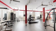 Located at 1901 S Charles St in Baltimore's South Baltimore neighborhood, this fitness center offers a thoughtfully designed, single-floor layout that accommodates a comprehensive workout experience. The main gym area features durable rubber flooring and is generously proportioned, providing ample space for a wide array of cardio and strength equipment, including treadmills, elliptical machines, stationary bikes, cable machines, and a multi-tiered dumbbell rack. Bold red structural columns and matching accent stripes along the upper walls create a dynamic, energizing atmosphere against a neutral cream backdrop. Track lighting and ceiling fans run throughout, ensuring the space remains well-lit and ventilated. Large windows fitted with horizontal blinds line the main workout area, inviting natural light while maintaining privacy. A dedicated studio room features warm hardwood flooring, floor-to-ceiling mirrors framed in red, and a rack of medicine balls and exercise balls, making it ideal for stretching, yoga, or functional training. An entry foyer with additional hardwood flooring and a wall-mounted resistance accessory organizer provides a smooth transition into the gym. The overall design is clean, modern, and functional, delivering a professional fitness environment well-suited for a variety of training needs.