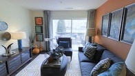This single-floor space showcases a thoughtfully designed living area that blends contemporary style with warm, inviting accents. The living room serves as the focal point, featuring a large sliding glass door that floods the space with natural light and opens onto a private balcony, offering pleasant outdoor views. The interior is distinguished by a striking burnt orange accent wall that adds depth and warmth to the room, complemented by neutral cream walls and patterned drapes that frame the expansive window. The living area is furnished with a plush charcoal sectional sofa and matching armchair, accented with bold geometric and animal-print throw pillows that add a dynamic visual layer. A gold-and-white geometric area rug anchors the seating arrangement, while a mirrored tray coffee table and round ottoman contribute to the refined, curated aesthetic. Decorative touches such as framed wall art in warm tones, sculptural vases, and potted greenery enhance the room's personality. The hard flooring adds a clean, modern foundation throughout. Overall, the space presents a well-balanced combination of bold color choices, layered textures, and purposeful décor, creating a warm and inviting atmosphere with seamless indoor-outdoor connectivity.