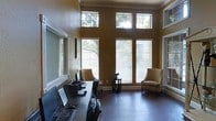 This single-floor space makes a strong impression with its dramatic ceiling height and expansive windows that flood the interior with natural light. The main area serves as a well-appointed office, where a long, dark-toned work desk lines one wall and is equipped with multiple computer workstations, making it ideal for focused productivity. Warm golden-hued walls contribute to an inviting and professional atmosphere throughout.

A row of large windows fitted with horizontal blinds lines the front wall, allowing for adjustable light control while maintaining a connection to the lush outdoor greenery visible beyond. Above these windows, a second tier of transom windows draws additional daylight deep into the room, enhancing the sense of openness and vertical scale.

Complementing the workspace, a comfortable seating area occupies the far end of the room, featuring two upholstered armchairs in a neutral tone arranged around a small side table — ideal for informal meetings or relaxed conversation. Dark hardwood flooring runs throughout, grounding the space with richness and contrast against the warm wall color. Crown molding adds a refined architectural detail, reinforcing the polished and professional character of the overall environment.