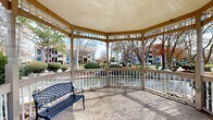 This gazebo in Kenner, Louisiana, is a well-appointed open-air structure designed to serve as a relaxing retreat within a landscaped community setting. The octagonal design features a solid canopy roof supported by decorative columns, with ornamental lattice detailing along the upper perimeter that adds a classic architectural character. A white-painted balustrade railing encloses the perimeter, providing a sense of enclosure while maintaining unobstructed views of the surrounding grounds.

The flooring consists of a mixed brick and gravel surface, lending a traditional, finished aesthetic underfoot. A wrought-iron bench provides seating within the structure, making it an ideal spot for relaxation. The gazebo is positioned alongside a water feature, offering pleasant waterfront views from multiple vantage points. Mature trees and manicured shrubs frame the exterior, enhancing the natural ambiance.

From within the gazebo, views extend to a community swimming pool surrounded by a brick paver deck furnished with lounge chairs and picnic tables, ideal for outdoor leisure. The surrounding multi-story residential buildings are visible in the backdrop, set against an open sky. The overall setting combines functional outdoor design with a serene, park-like atmosphere.