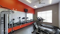 This fitness center is thoughtfully designed to provide a functional and motivating workout environment. The space is defined by bold red accent walls paired with a neutral gray floor, creating an energetic atmosphere that encourages activity. A full-length mirrored wall lines one side of the room, visually expanding the space while allowing users to monitor their form during exercise.

The facility is outfitted with a variety of cardio equipment, including treadmills, elliptical machines, and a stationary bike, all arranged to maximize floor space and usability. A free weight station with a dumbbell rack is also visible, catering to those seeking strength training options. Overhead, a ceiling fan provides air circulation throughout the workout area, complementing the bright fluorescent lighting for a well-lit environment.

A wall-mounted flat-screen television is positioned prominently for viewing during workouts, adding an entertainment element to the space. Natural light filters in through a large window fitted with blinds, offering a connection to the outdoors. The textured ceiling adds a classic architectural touch, while the overall layout of the room ensures a comfortable and efficient space for a full range of fitness activities.