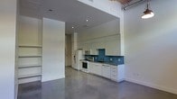 This single-floor space showcases a contemporary, industrial-inspired design that seamlessly blends open-concept living with functional details. The expansive main area is anchored by polished concrete floors that extend throughout, lending a clean, modern aesthetic while reflecting the abundant recessed lighting overhead.

The ceilings soar to impressive heights, with exposed ductwork visible in portions of the space, reinforcing the loft-like character of the unit. Crisp white walls throughout create a bright, airy atmosphere that enhances the sense of openness.

The kitchen is a standout feature, outfitted with white flat-front cabinetry that extends to the ceiling, offering generous storage. A vibrant teal-blue tile backsplash adds a bold pop of color and visual interest. Dark countertops provide a striking contrast, while stainless steel appliances — including a gas range, microwave, refrigerator, and dishwasher — complete the well-equipped cooking space.

Adjacent to the main living area, a recessed nook with open built-in shelving offers flexible storage or display space. Structural columns and thoughtfully placed archways define zones within the open floor plan without sacrificing the sense of flow. Overall, this space delivers a refined urban aesthetic with practical, well-considered features throughout.